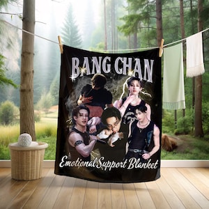 May include: Black blanket featuring the text "BANG CHAN" in white, with images of a male musical artist. The words "Emotional Support Blanket" are printed below the images. The blanket is hanging on a clothesline.