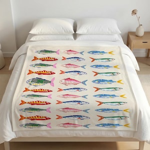 May include: A cream-colored blanket featuring a repeating pattern of colorful fish illustrations. The fish are in various colors, including green, red, blue, and yellow, with pink or yellow fins. The blanket is on a bed with white bedding.
