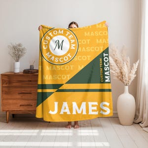 Pode incluir: Um cobertor amarelo e verde com o nome "JAMES" em letras brancas. O cobertor apresenta um logotipo circular com o texto "CUSTOM TEAM MASCOT" e a letra "M".