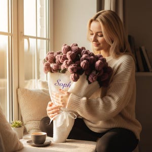 May include: A bouquet of crocheted flowers in shades of mauve and purple, wrapped in white fabric with the name "Soph" in script. The bouquet is tied with a white ribbon. A cup of coffee sits on a saucer nearby.