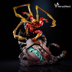 May include: A red and gold action figure of a superhero in a dynamic pose, with golden mechanical spider-like arms. The figure is perched atop a detailed, weathered base with a glowing green core. The VersaNest Studio logo is in the top right corner.