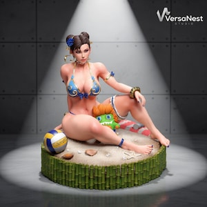Puede incluir: Una figura coleccionable de un personaje con cabello oscuro en moños, vistiendo un bikini azul y dorado, sentada sobre una base de arena rodeada de tallos de bambú. La figura está acompañada por una pequeña criatura de pelo naranja, un balón de voleibol y una estrella de mar.