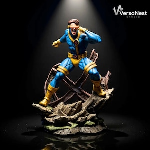 Op de afbeelding: Een gedetailleerd beeldje van Cyclops, een superheld, in een dynamische pose. Het beeldje draagt een blauw en geel kostuum, met een rood vizier. De basis is een gedetailleerd diorama van een rotsachtig terrein. Het VersaNest Studio logo is zichtbaar.