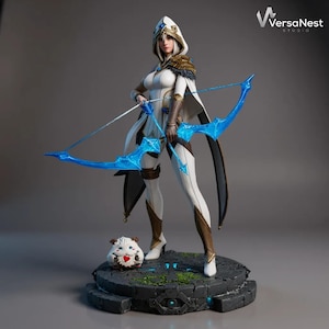 Puede incluir: Figura detallada de una arquera con un atuendo blanco y dorado, sosteniendo un arco y una flecha azules. La figura está sobre una base de piedra con una pequeña criatura. La figura tiene una capucha y una capa. El logotipo de VersaNest Studio está en la esquina superior derecha.