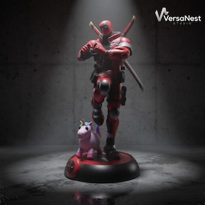 Puede incluir: Una figura de Deadpool roja y negra de pie sobre una base circular con un pequeño unicornio rosa. El personaje lleva una máscara, un cinturón de herramientas y dos espadas en la espalda. La base presenta un logo de Deadpool. El texto VersaNest STUDIO es visible.