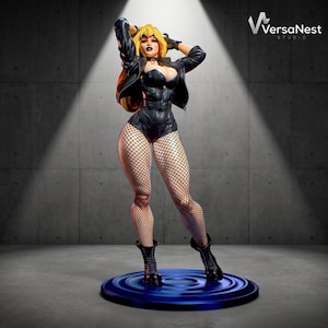 Puede incluir: Una figura coleccionable de un personaje rubio con chaqueta de cuero negra, body negro y medias de rejilla negras. El personaje tiene labios oscuros y está de pie sobre una base circular azul. El logotipo de VersaNest Studio es visible.