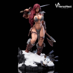 May include: A detailed figurine of a warrior woman with red hair, clad in armor, holding a sword and axe. The figure stands on a snowy base with a shield and arrows, against a black backdrop. The VersaNest Studio logo is visible.