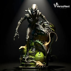 May include: A detailed sculpture of an Alien creature, posed menacingly. The figure is metallic silver with sharp teeth and claws, standing on a base with alien eggs and green, organic elements. The VersaNest Studio logo is visible.