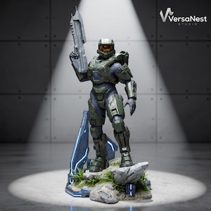 Op de afbeelding: Een gedetailleerd Master Chief-figuur op een rotsachtige basis met groen gras en blauwe lichtaccenten. Het personage draagt een olijfgroen harnas met een gouden vizier en houdt een grijs geweer omhoog in de rechterhand. Het VersaNest Studio logo is zichtbaar.