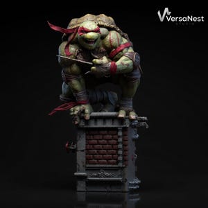Op de afbeelding: Een gedetailleerd beeld van Raphael van Teenage Mutant Ninja Turtles, geplaatst op een bakstenen structuur. De figuur is groen met een rood masker en houdt een sai vast. Het VersaNest Studio-logo is rechtsboven zichtbaar.