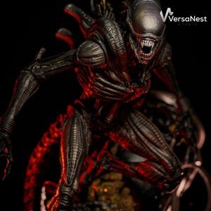 Puede incluir: Una figura detallada y oscura de Alien en una pose amenazante con la boca abierta. La figura tiene una apariencia texturizada y met&aacute;lica, con dientes visibles y una cabeza elegante y alargada. El fondo es oscuro, con iluminaci&oacute;n roja que a&ntilde;ade al efecto dram&aacute;tico. El logo de VersaNest Studio es visible.
