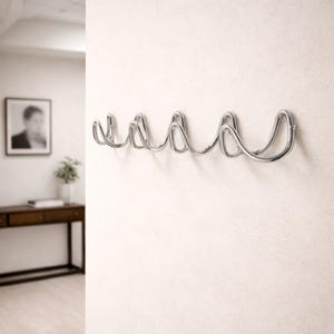 Può includere: Un attaccapanni da parete moderno con finitura cromata. Presenta un design ondulato continuo con più ganci per appendere gli indumenti. È montato su una parete bianca, con un quadro e un tavolino visibili sullo sfondo.