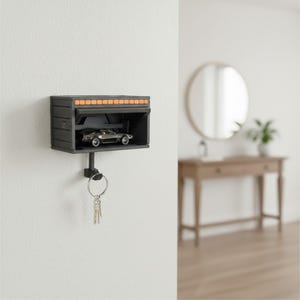 Könnte beinhalten: Ein schwarzer, an der Wand befestigter Schlüsselhalter in Form einer Miniaturgarage mit einem orangefarbenen Lichtstreifen über dem Garagentor. Ein kleines silbernes Auto befindet sich im Inneren. Schlüssel hängen darunter.