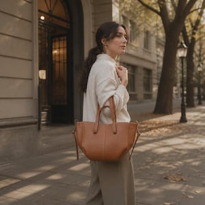 Peut inclure: Un sac cabas en cuir marron clair avec deux anses supérieures et des détails de nœuds sur les côtés. Le sac présente une base structurée et incurvée avec une finition lisse. Il est porté à l'épaule, affichant un design épuré avec des coutures.