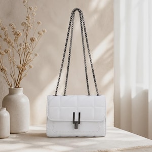 Peut inclure: Un sac à bandoulière blanc matelassé présentant une silhouette rectangulaire avec un motif à carreaux. Le sac comprend une bandoulière en chaîne métallique foncée et un fermoir rectangulaire assorti sur le rabat avant pour la fermeture.