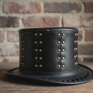May include: Black top hat with brass-colored studs. The hat has a wide brim and a cylindrical crown. The hat is made of a leather-like material and is likely a costume accessory or a fashion statement.