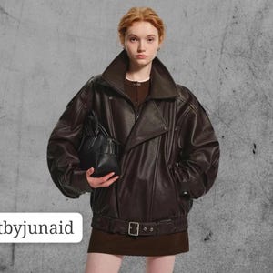 Peut inclure: Veste en cuir marron foncé avec un large col et une ceinture à la taille. La veste a une fermeture éclair et un petit sac à main noir. Le texte "craftbyjunaid" est visible.