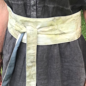 May include: A wide, light green fabric belt with a blue and white stripe on one end. The belt is tied around a person's waist and is knotted in the front.