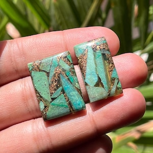 Puede incluir: Dos cabujones rectangulares de piedras preciosas con un mosaico de inclusiones turquesas y color bronce. Las piedras se sostienen en una mano, mostrando sus superficies pulidas y patrones únicos. Los cabujones miden aproximadamente 2.5 cm de largo.