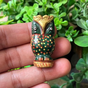 May include: A small, hand-painted wooden owl figurine. The owl has a green body with red and yellow polka dots, a gold-colored head, and dark green eyes. The figurine is held in a hand against a background of green foliage.