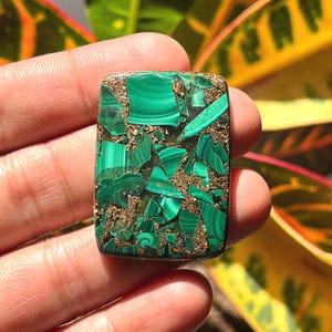 May include: Rectangular malachite stone with a mosaic pattern of deep green and gold-brown fragments. The polished surface reflects light, showcasing the stone's natural textures and colors. This gemstone is ideal for jewelry making or collecting.