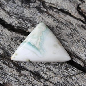 Puede incluir: Una gema triangular con una base blanca y patrones arremolinados de azul pálido y verde. La piedra pulida se encuentra sobre una superficie de madera gris desgastada, resaltando su belleza natural. Esta piedra única podría usarse para joyería u otras manualidades.