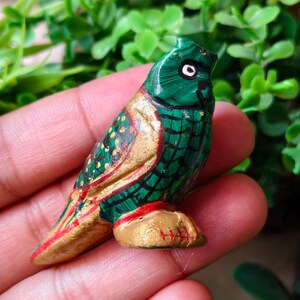 May include: A hand-painted wooden owl figurine. The owl is primarily green with gold and red accents. The owl's body is green with a scale-like pattern, and the wings and base are gold. The owl has a white eye and a black pupil.