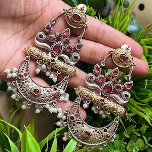 Op de afbeelding: Oorbellen in zilver- en goudtinten met robijnrode edelstenen en kleine witte parelaccenten. De oorbellen hebben een halve maanvorm met ingewikkeld filigraanwerk en een parelkwastje. De oorbellen zijn ontworpen voor doorboorde oren.