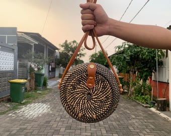 May include: A round, woven rattan handbag with a spiral design in black and tan. The bag features brown leather straps and a leather closure. The bag is held up by a person's hand.