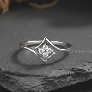 May include: A silver ring with a four-petal flower design set with small, clear gemstones. The ring has a delicate, curved band and a geometric, diamond-shaped setting. The ring is displayed on a dark, textured surface.