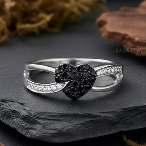 May include: A silver ring featuring a heart-shaped cluster of black stones. The band splits into two, lined with small white stones, and curves to embrace the heart. The ring is displayed on a dark, textured surface.
