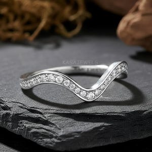 May include: A silver-colored ring with a V-shaped design. The ring is encrusted with small, clear gemstones along the top curve. The ring sits on a dark gray, textured surface. The brand name "KASA JEWELS" is visible.