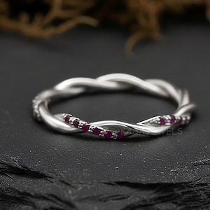 May include: A silver ring with a twisted design. Small, red gemstones are set into the ring. The ring is displayed on a black surface, with a blurred background of brown and green.