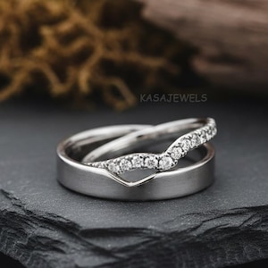 May include: Two silver rings, one with a V-shaped design and the other with a row of small, sparkling stones. The rings are stacked on a dark, textured surface, with a blurred background of moss and wood.