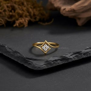 May include: A gold-colored ring with a diamond-shaped setting holding four small, clear gemstones. The ring has a delicate, curved band and is displayed on a dark, textured surface. The brand name "KASAJewels" is visible.