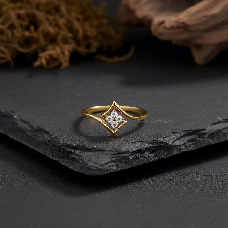 May include: A gold-colored ring with a diamond-shaped setting holding four small, clear gemstones. The ring has a delicate, curved band and is displayed on a dark, textured surface. The brand name "KASAJewels" is visible.