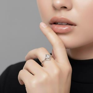 May include: A silver ring with a diamond-shaped setting featuring four small, clear gemstones. The ring is on a finger, with the hand and part of a person's face visible. The ring is simple and elegant.