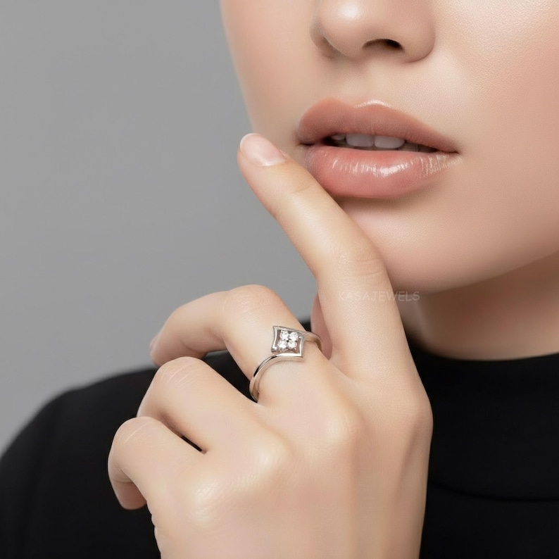 May include: A silver ring with a diamond-shaped setting featuring four small, clear gemstones. The ring is on a finger, with the hand and part of a person's face visible. The ring is simple and elegant.