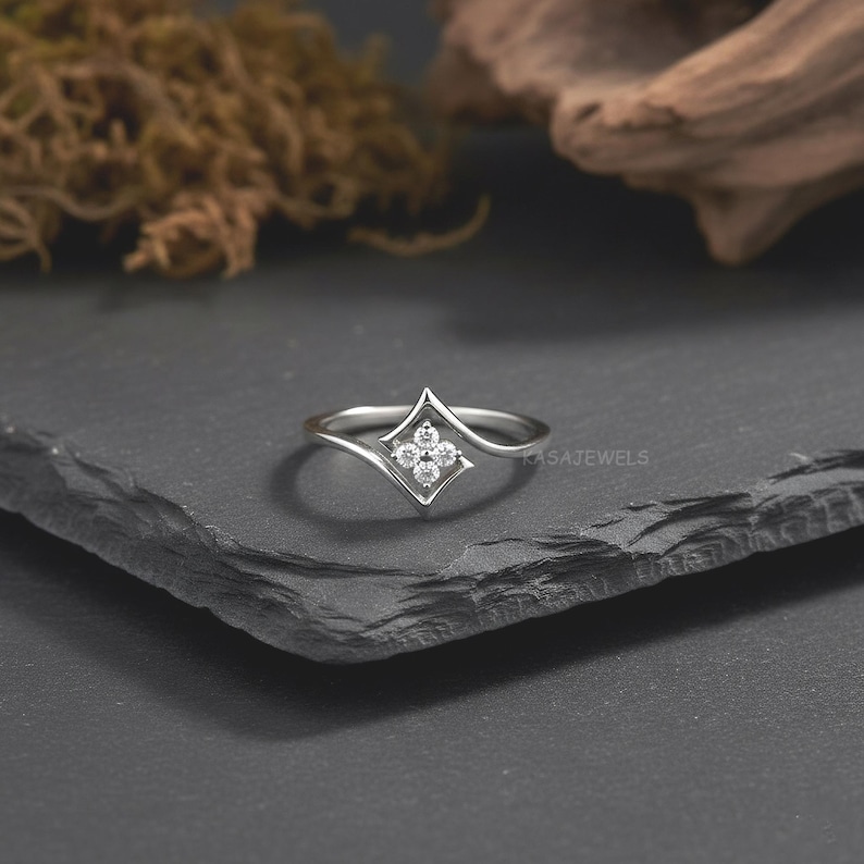 May include: A silver ring with a four-petal flower design set with small, clear gemstones. The ring has a delicate, curved band and a geometric, diamond-shaped setting. The ring is displayed on a dark, textured surface.