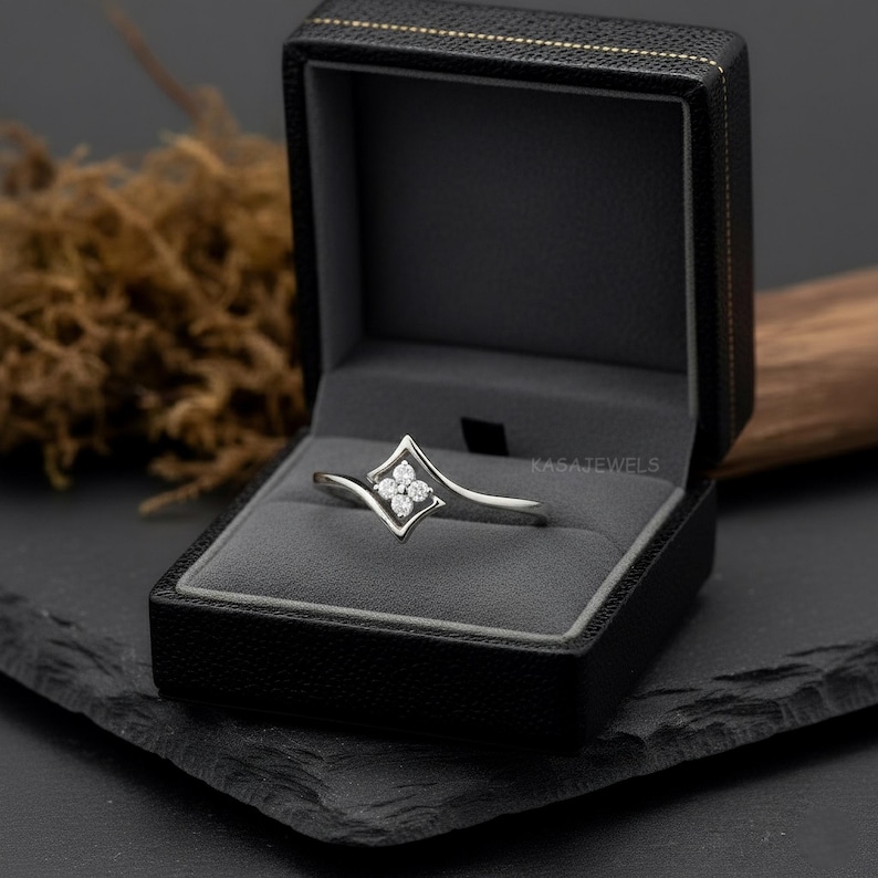 May include: A silver ring with a diamond-shaped centerpiece containing four small diamonds, presented in a black jewelry box. The ring has a curved design and is set against a dark background.