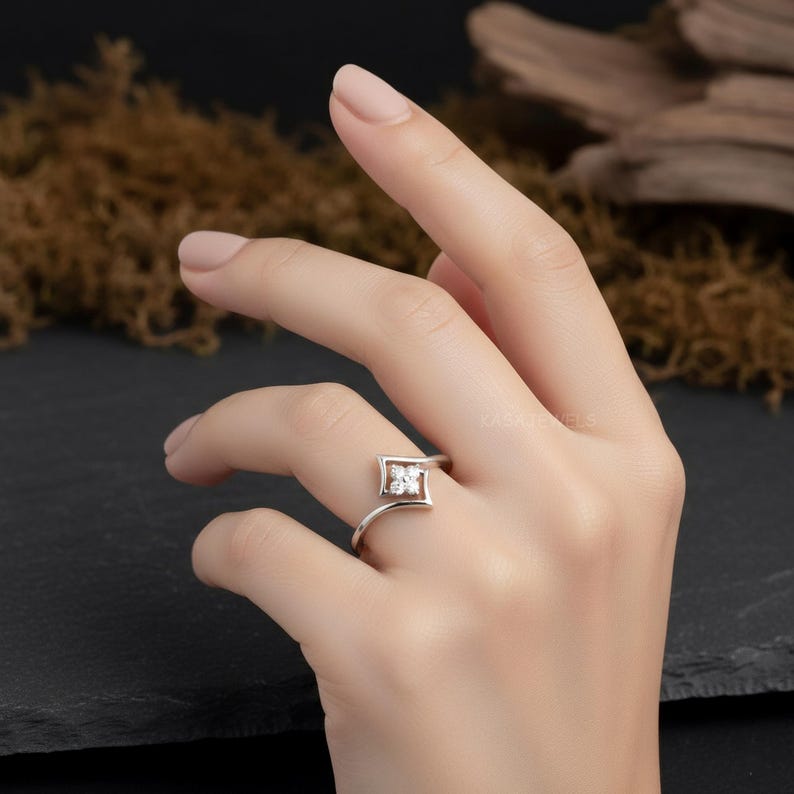 May include: A silver ring featuring a square-shaped design with a cluster of small, clear gemstones at the center. The ring's band curves gracefully, and the design is set against a neutral background.