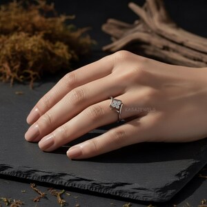 May include: A silver ring with a square, floral-inspired design. The ring features a central cluster of small, clear stones, set within a square frame. The ring is on a finger, with the brand name "KASAJEWELS" visible.