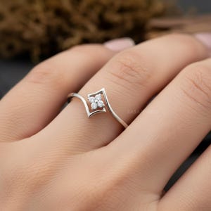 May include: A delicate silver ring featuring a square-shaped design with four small, round, clear gemstones. The ring has a unique, open design that curves around the finger. The ring is shown on a person's finger.