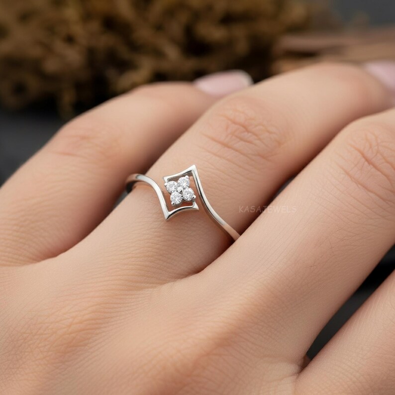 May include: A delicate silver ring featuring a square-shaped design with four small, round, clear gemstones. The ring has a unique, open design that curves around the finger. The ring is shown on a person's finger.
