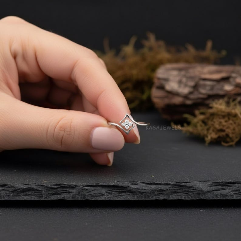 May include: A silver ring with a diamond-shaped center, featuring four small, clear gemstones. The ring's band curves gracefully, and the design is held by a hand against a dark background. The ring is a piece of jewelry.