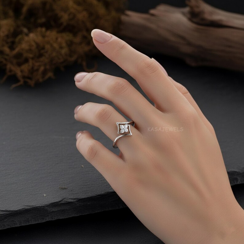 May include: A silver ring with a square-shaped setting featuring four small, clear gemstones. The ring's band curves gracefully, and the brand name "KASAJEWELS" is visible in the background. The ring is displayed on a hand.