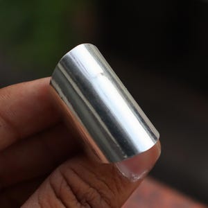 May include: A polished silver metal ring, cylindrical in form. The ring's surface is smooth and reflects light. The ring is about 2.5 cm long and is likely a finger ring.