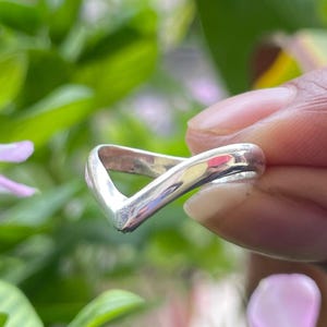 May include: A silver-colored ring with a V-shaped design. The ring is smooth and polished, reflecting light. The ring is held in front of a background of green leaves and pink flowers.