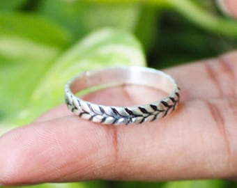 May include: A silver ring with a leaf pattern. The ring has a repeating design of stylized leaves around the band. The ring is made of a shiny metal. The background is a blurred green.