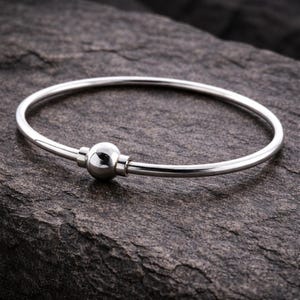 Könnte beinhalten: Ein silberfarbenes Armreif-Armband mit einem kugelförmigen Verschluss. Das Armband ist kreisförmig und hat eine glatte, polierte Oberfläche. Der Verschluss ist ein kleiner, runder Ball, der von zwei zylindrischen Teilen gehalten wird.